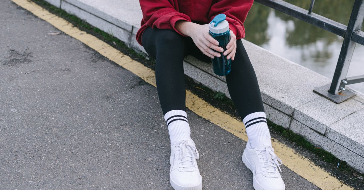 Person sitting on a street curb holding a sports water bottle, wearing sneakers.