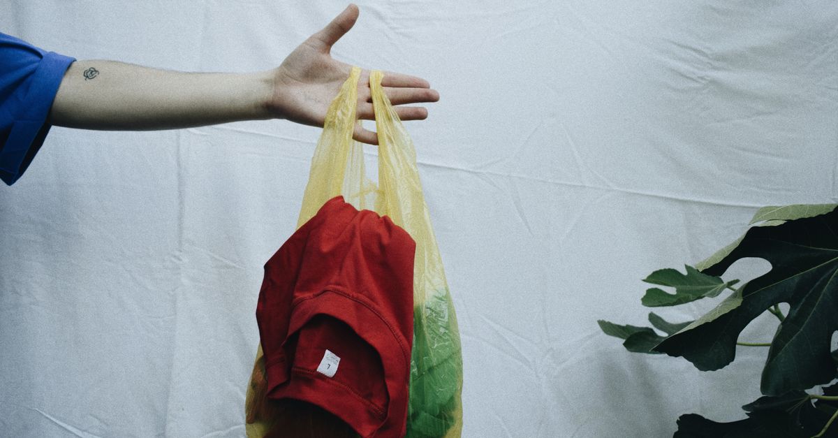 A hand holds a yellow plastic bag containing clothes on a simple white background.
