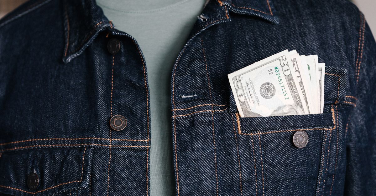 Crop unrecognizable male in casual outfit standing with different nominal pars of dollar banknotes in pocket of jeans jacket