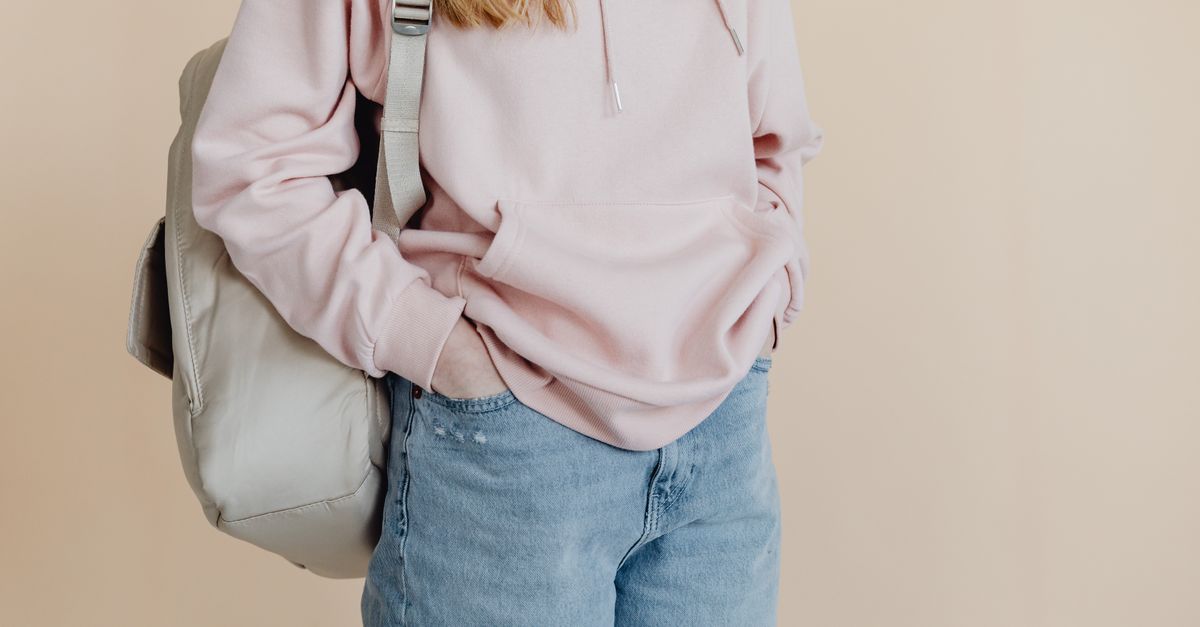 A teenager in a casual pink hoodie and jeans carrying a backpack, standing against a neutral background.